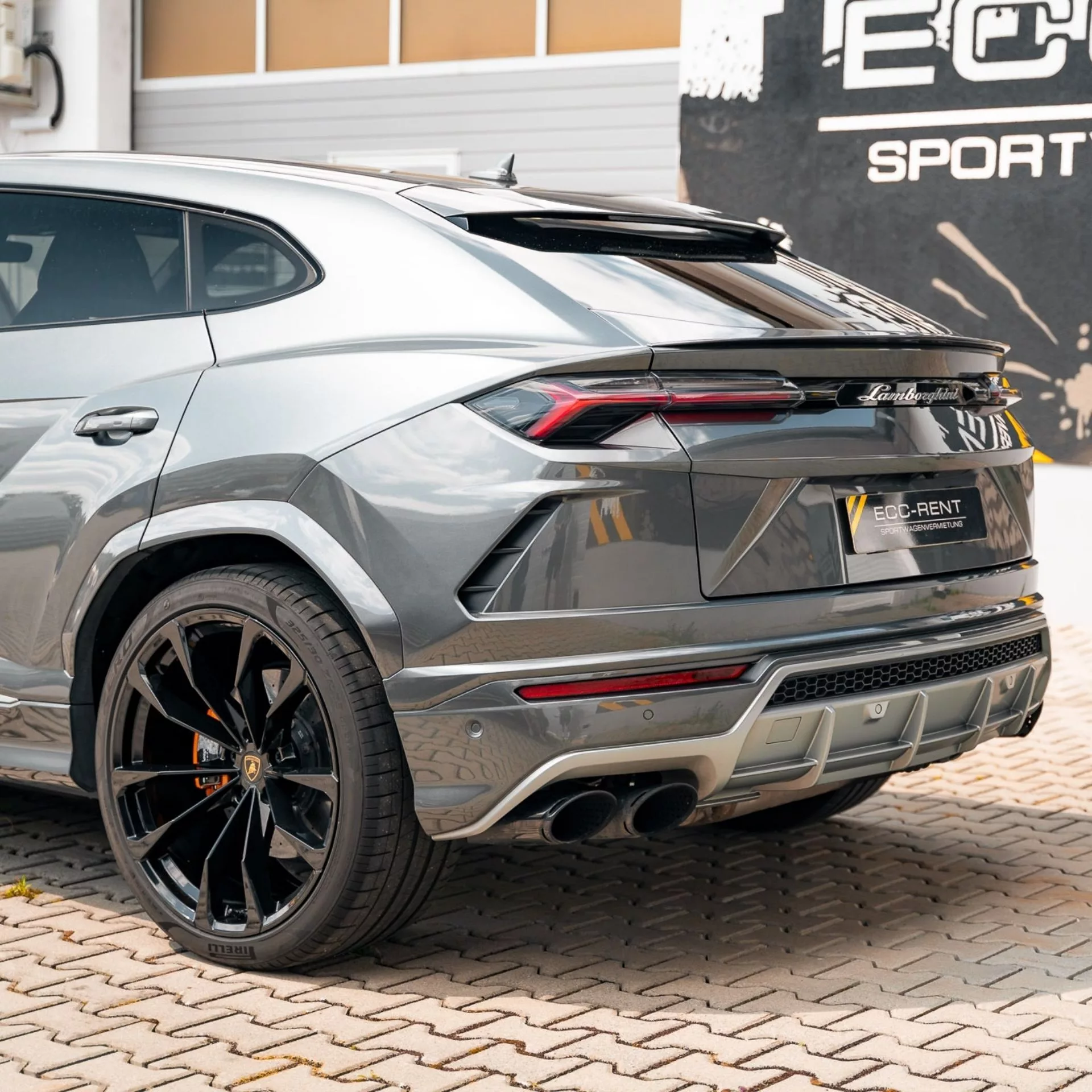 Grauer Lamborghini Urus auf gepflastertem Parkplatz vor einem Gebäude.